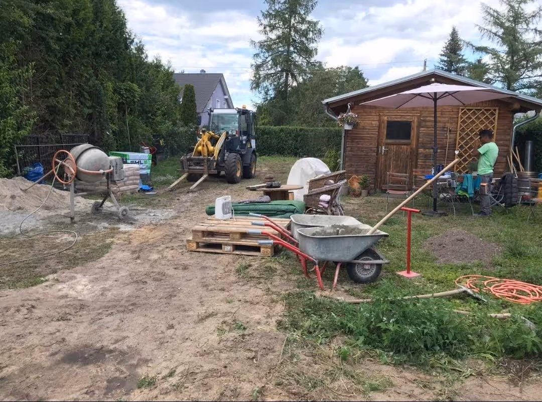 Baustelle im Garten mit Radlader, Betonmischer und Materialpaletten vor dem Gartenhaus