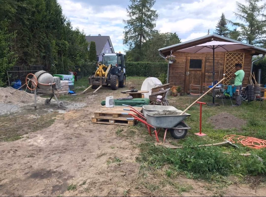 Baustelle im Garten mit Radlader, Betonmischer und Materialpaletten vor dem Gartenhaus