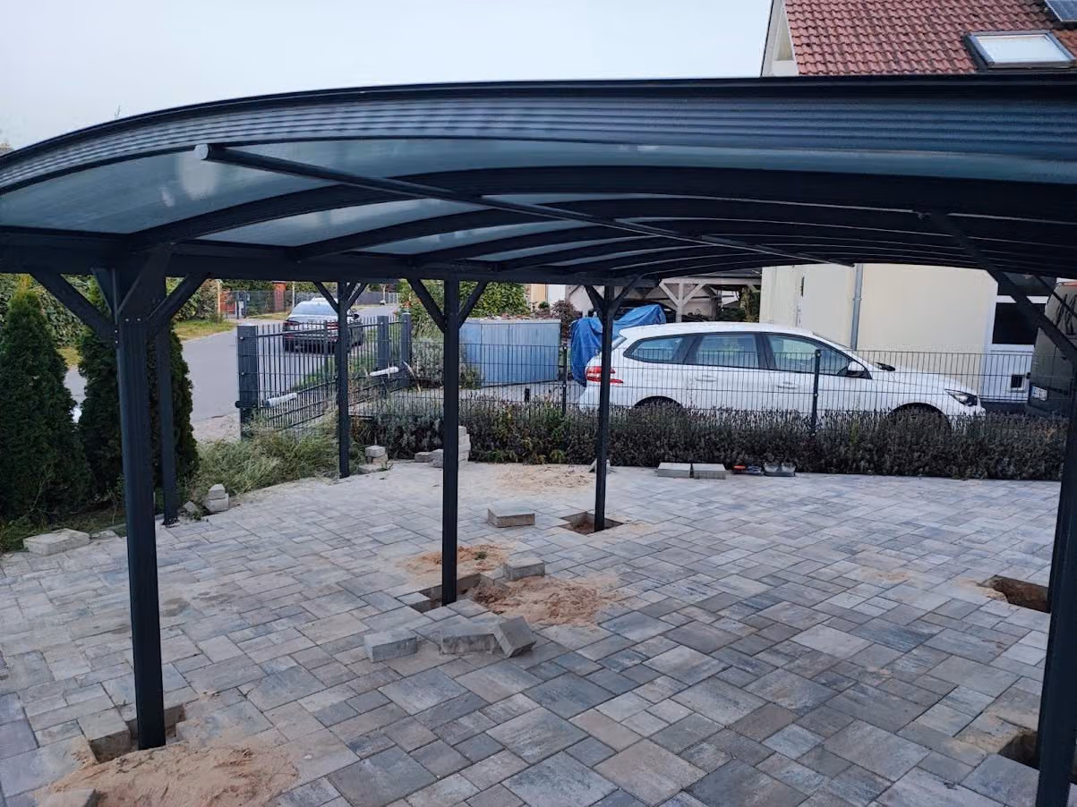 Carport mit Bogendach auf einer gepflasterten Stellplatzfläche, Stützen im Fundament