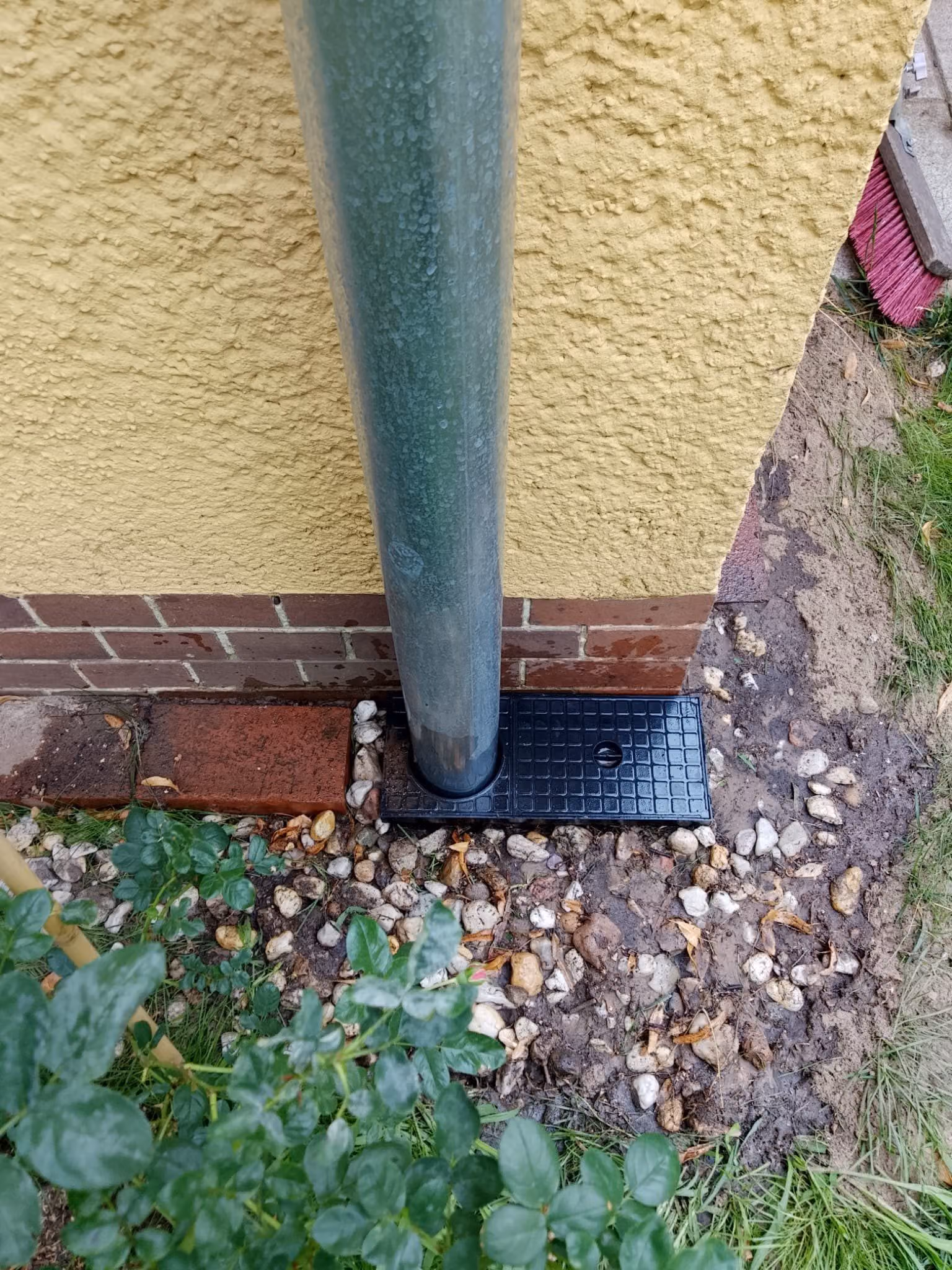 Regenfallrohr endet über einem Sinkkasten mit Gussrost, ringsum ein Kiesstreifen am Sockel