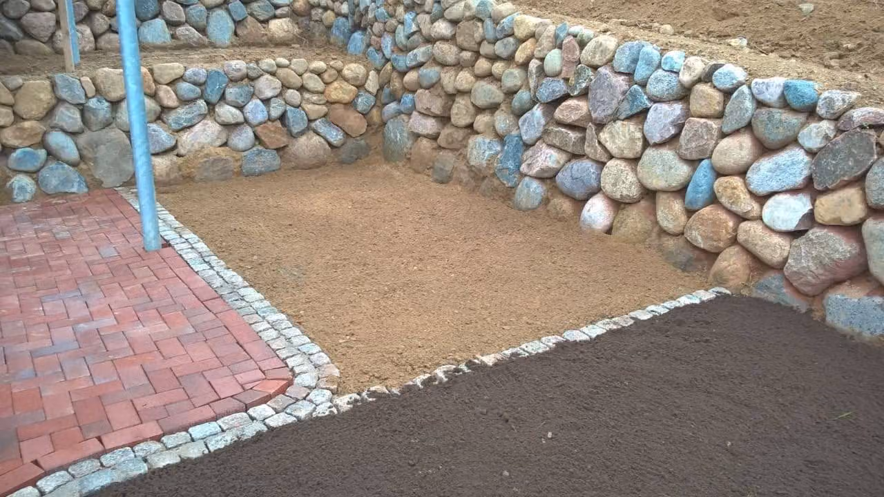 Zweistufige Feldsteinmauer aus runden Lesesteinen, davor ein Weg aus rotem Klinkerpflaster mit Granit-Kleinpflasterkante