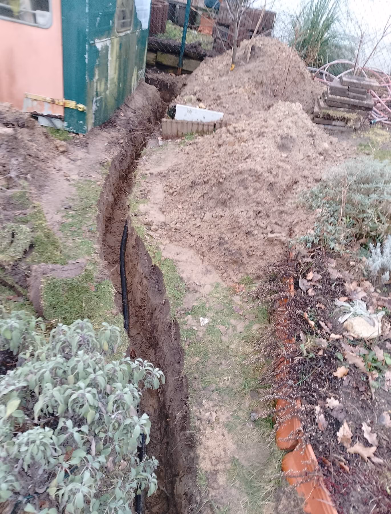 Schmaler Kabelgraben entlang des Gartenschuppens, das schwarze Erdkabel liegt auf der Sohle, Aushub daneben