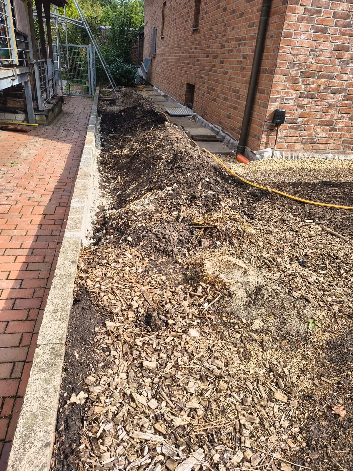 Offener Arbeitsgraben an der Hauswand mit freigelegtem Klinkersockel, daneben der Gartenweg