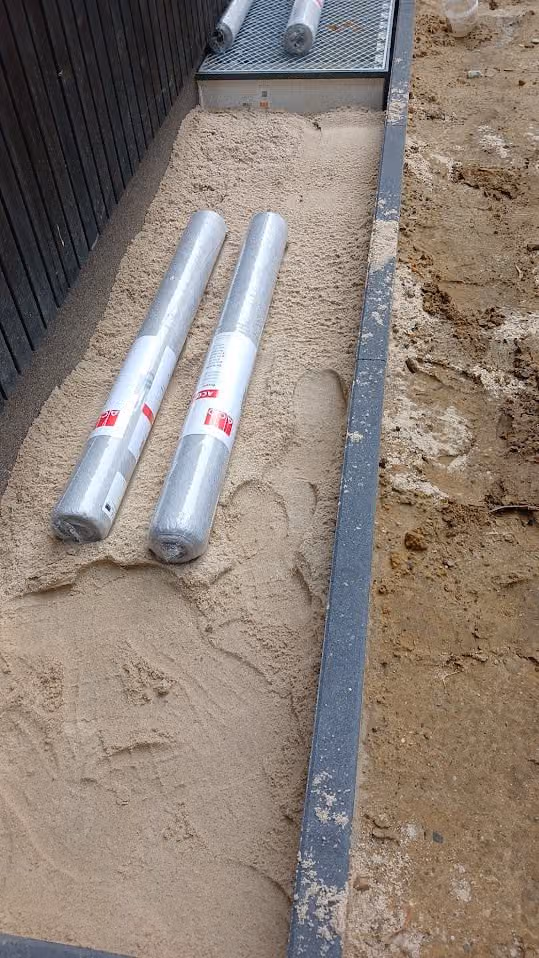 Zwei Rollen Vlies liegen im Sandbett des künftigen Spritzschutzstreifens