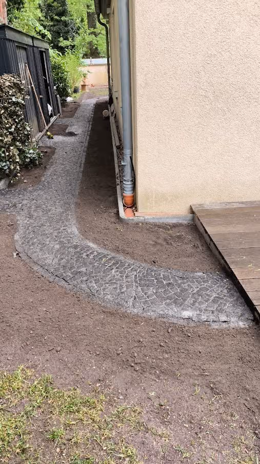 Geschwungener Hausumgang aus Mosaikpflaster entlang der Fassade, Erdreich noch offen