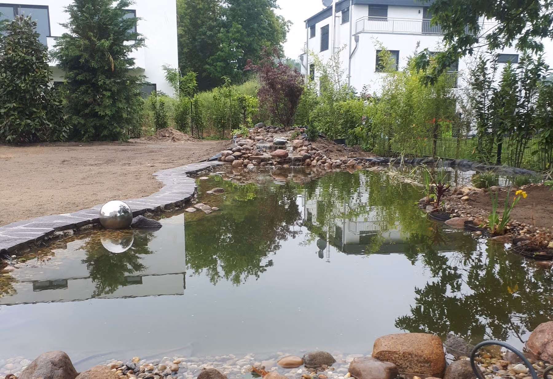 Fertiger Naturteich mit spiegelnder Wasserfläche, Bachlauf im Hintergrund, Kantensteinen an der Uferlinie und einer Flachwasserzone aus Kies und Findlingen