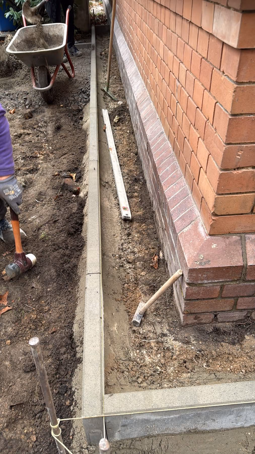 Betonrandstein steht im Betonbett entlang der Klinkerfassade, an der Gebäudeecke auf Gehrung geschnitten, Wasserwaage und Schnur liegen an