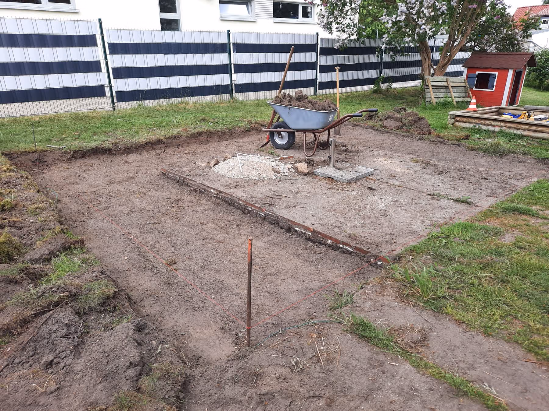 Ausgehobene Terrassenfläche mit gespannter Schnur und Eisenpfosten als Höhenmaß, Schubkarre mit Aushub daneben